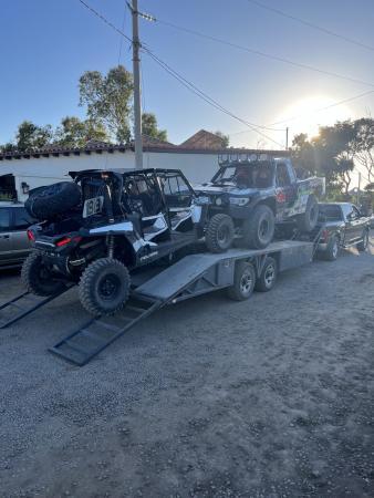 A black and white off-road vehicle and a trophy truck loaded onto a flatbed trailer attached to a pickup truck.