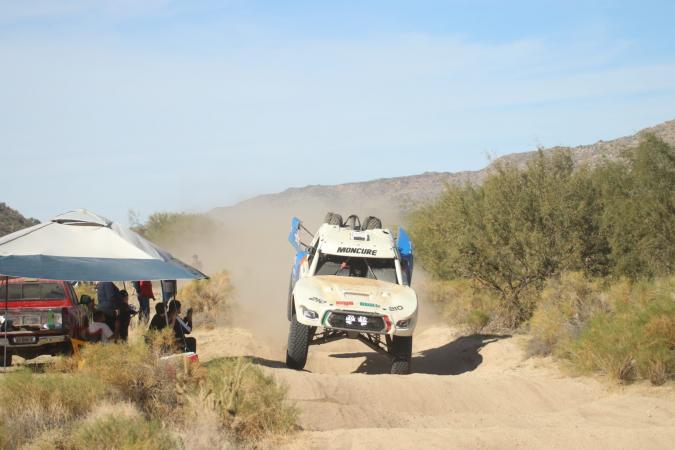 A white off-road racing truck speeds along a desert dirt track, kicking up dust near a group of spectators gathered under a canopy.