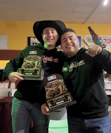 Two men smiling and holding first-place trophies for the 2025 off-road championship, with one man holding a beer bottle.