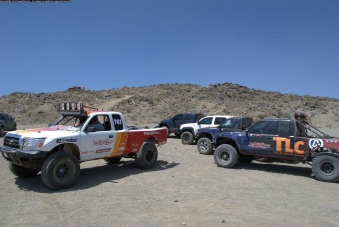 Three off-road racing trucks parked on a dirt trail in a desert landscape.