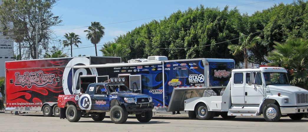 A white semi-truck pulls a large, branded race trailer parked next to a blue and red Long Beach Racers Toyota trophy truck.