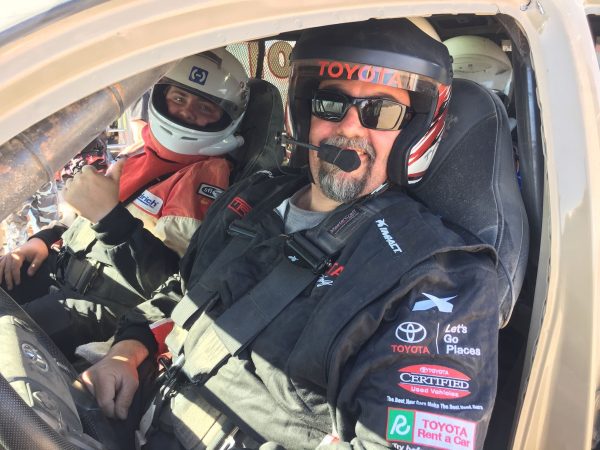 Two people wearing racing helmets and fire suits sit inside a vehicle, with the driver in the foreground giving a thumbs-up.