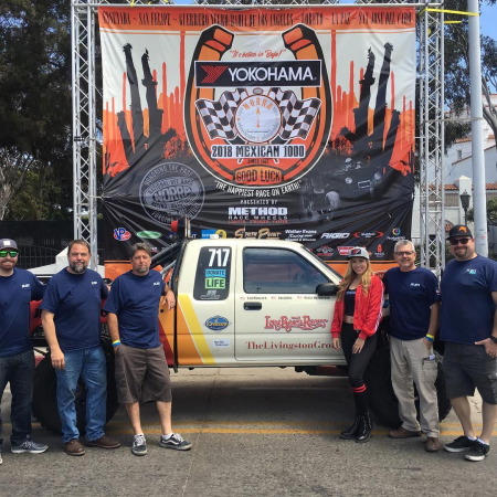 A group of six people posing in front of a NORRA Mexican 1000 race truck, with a large event banner hanging on a metal frame in the background.