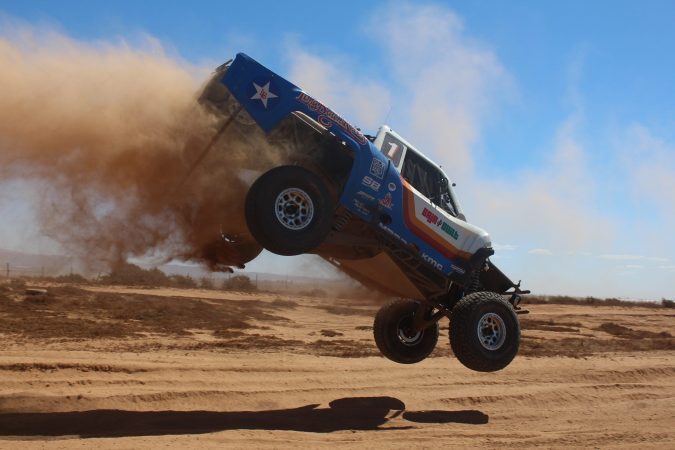 A blue and white trophy truck jumping over a dirt track, kicking up a large cloud of dust.