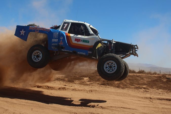 A blue and white trophy truck jumps through the air over a dirt track, kicking up a large cloud of dust.