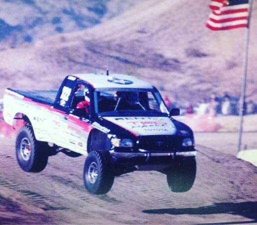 A Toyota off-road racing truck jumps over a dirt ramp with an American flag flying in the background.