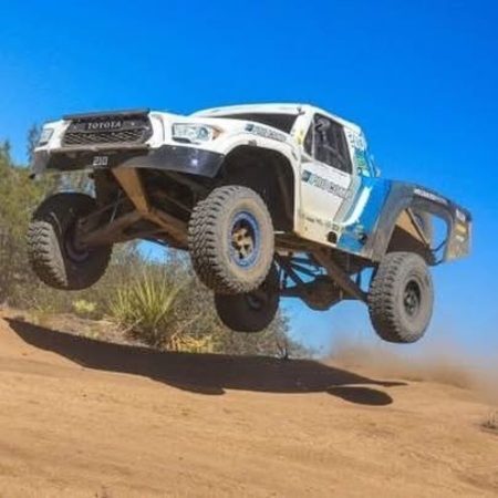 A white Toyota off-road racing truck jumps over a dirt mound against a clear blue sky.