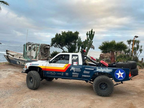 A white and blue off-road racing truck with number 717 parked next to an old, weathered boat on a cliffside overlooking the ocean.