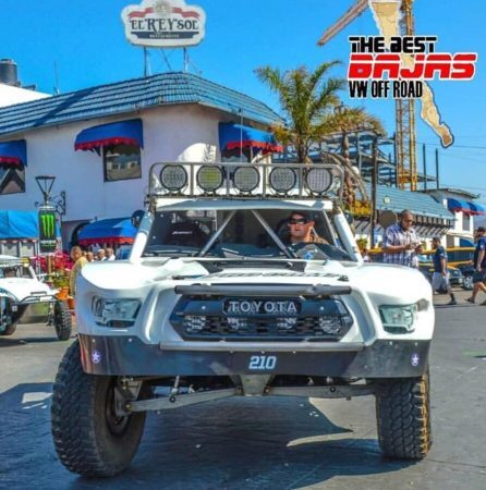 A white Toyota trophy truck with a light bar and the number 210 parked in front of a building with an El Rey Sol sign.