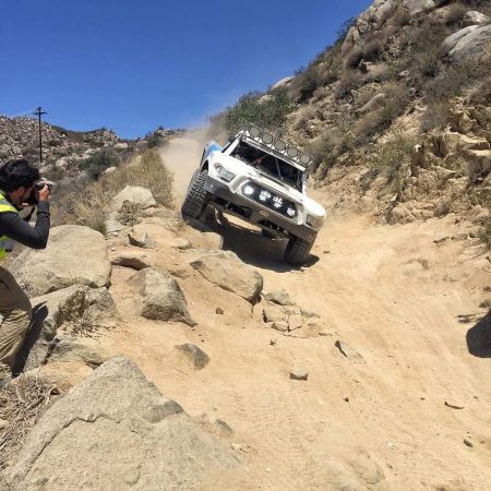 A photographer stands on a rocky hillside to take a picture of a white off-road racing truck climbing a steep, sandy trail.