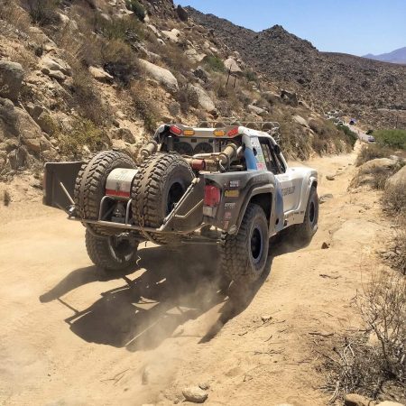 A trophy truck driving along a dusty, rocky desert trail with two spare tires mounted on the rear.