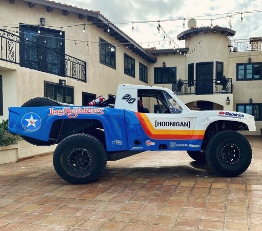 A Hoonigan off-road race truck with blue, white, and orange racing decals parked in front of a house.