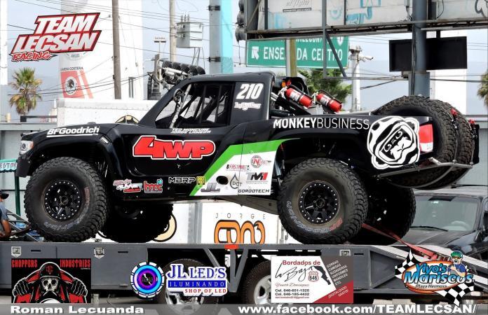 A black off-road racing truck numbered 210, labeled Monkey Business, secured on a flatbed trailer featuring various sponsor decals.