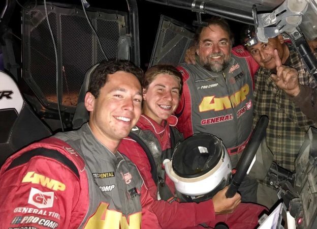 Four men posing inside a race vehicle, including two drivers in red and grey racing suits.