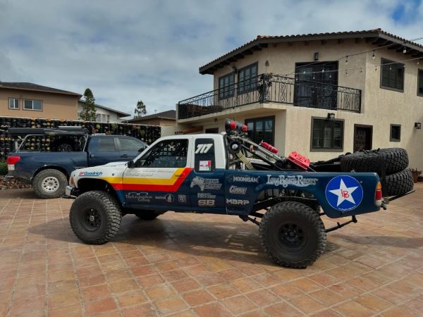 A modified off-road race truck with sponsor decals and a blue star logo parked on a residential patio next to a pickup truck.