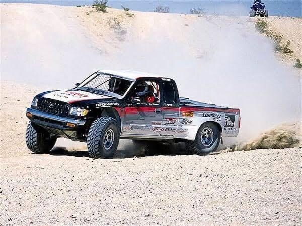 A Toyota off-road racing truck kicks up a cloud of dust while driving on a sandy track.