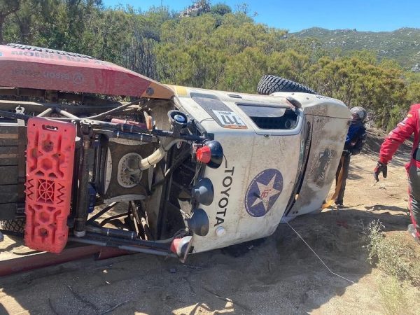 A white Toyota off-road racing truck overturned on its side on a dirt trail, with two people in racing gear standing nearby.