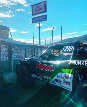 A black and green off-road race truck with the number 210 parked next to a building covered in stickers, beneath a Tecate and Acambaro business sign.