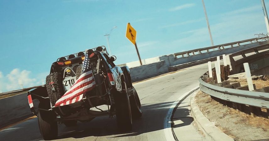 Off-road truck with a large American flag attached, driving on a curved highway ramp under a clear sky.