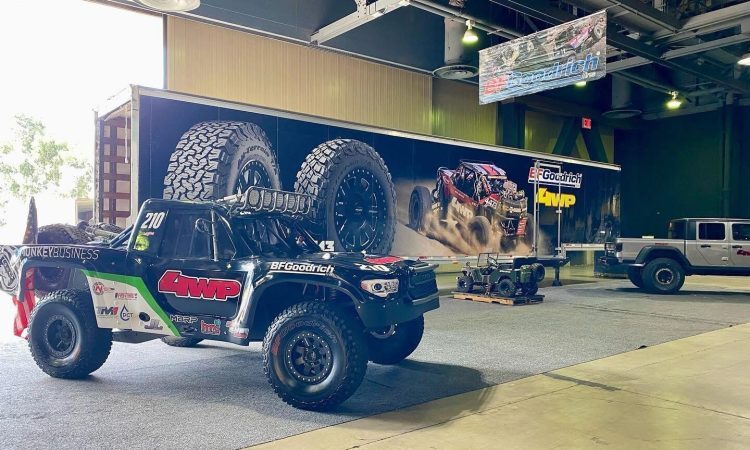 A black trophy truck numbered 210 with 4WP branding parked in front of a trailer featuring an image of an off-road racing vehicle.