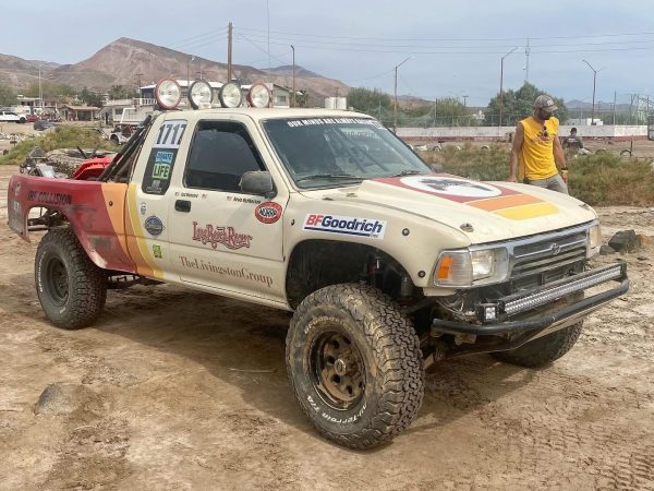 A white off-road racing truck with sponsor decals and the number 1717 parked on a dirt lot with mountains in the background.