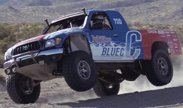 A blue and red Toyota off-road racing truck jumps over a dirt hill.