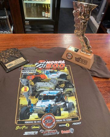 A brown t-shirt featuring a 2022 NORRA 500 graphic, displayed on a wooden table with a gold trophy and a square plaque.