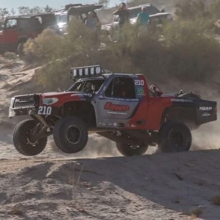 A red and grey trophy truck numbered 210 jumps over a dirt mound during an off-road race.