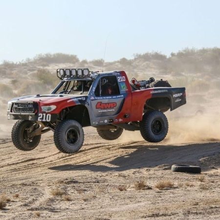 A red and grey Toyota trophy truck jumps over a dirt track in a desert landscape.