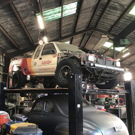 A race-prepped pickup truck elevated on a hydraulic lift above a vintage sedan in a workshop.