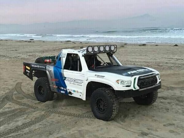 A white and blue Toyota trophy truck with off-road tires parked on a beach near the ocean.