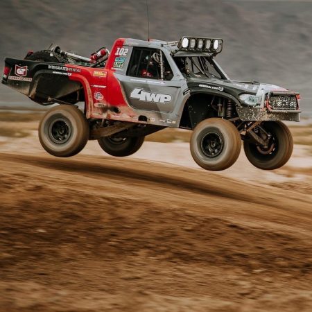 A trophy truck with red and grey livery jumping over a dirt mound.