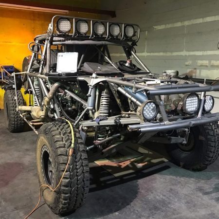 Off-road racing vehicle with a tubular steel frame, large tires, and multiple light bars, parked in a garage.