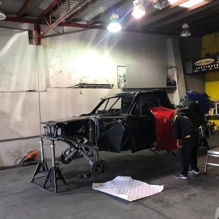 A mechanic in a garage works on the side of a black off-road race truck supported by jack stands.