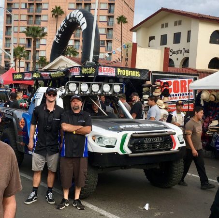 Two men stand in front of a modified Toyota off-road racing truck parked at an outdoor event.