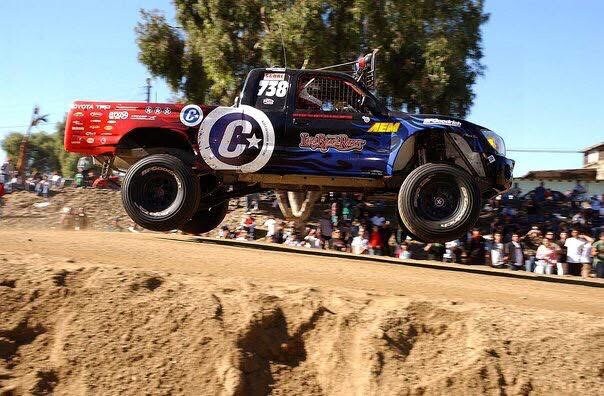 A race truck with the number 738 and a large C logo jumping over a dirt mound in front of a crowd.