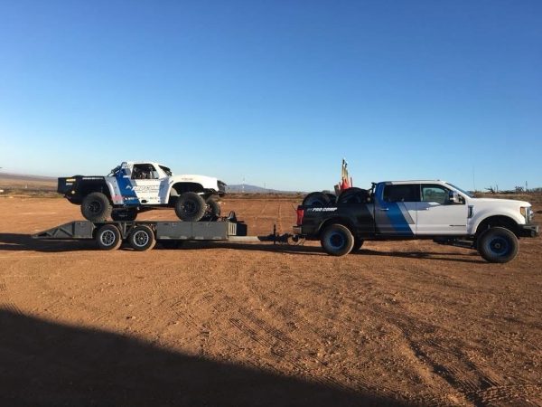 A white and black pickup truck towing a racing trophy truck on a flatbed trailer in a desert landscape.