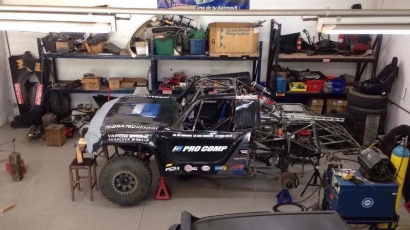 A trophy truck in a garage being assembled, with a bare chassis, off-road tires, and Pro Comp branding on the side.
