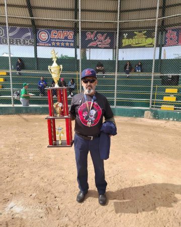 A man in a baseball cap, sunglasses, and team shirt stands on a dirt field holding a large three-tier trophy.