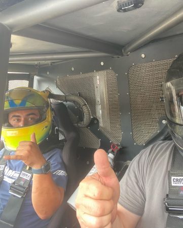 Two people in racing helmets inside a vehicle cockpit, one making a shaka sign and the other giving a thumbs up.
