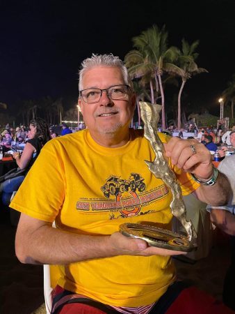 A smiling man in a yellow 2022 Yokohama NORRA Mexican 1000 t-shirt holding a metal trophy shaped like the Baja Peninsula.