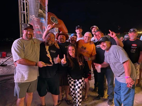 A group of people standing together at night, some holding drinks and one holding a racing trophy, in front of a race event banner.
