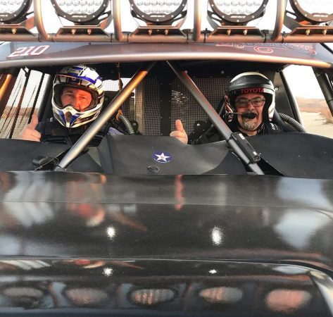 Two people in helmets sit inside a race car, both giving a thumbs-up gesture.
