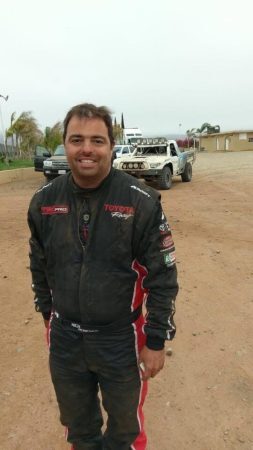 A race car driver in a black Toyota Racing suit stands in a dirt lot in front of a parked trophy truck.