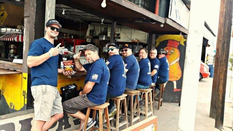 A group of six men in blue t-shirts sit on wooden stools at an outdoor bar counter.