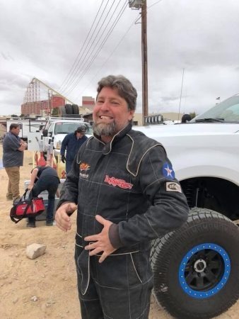 A smiling man in a black racing suit stands in front of a white off-road truck.