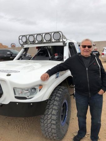 A man in a black zip-up hoodie and sunglasses stands next to a white off-road racing truck equipped with a roof-mounted light bar.