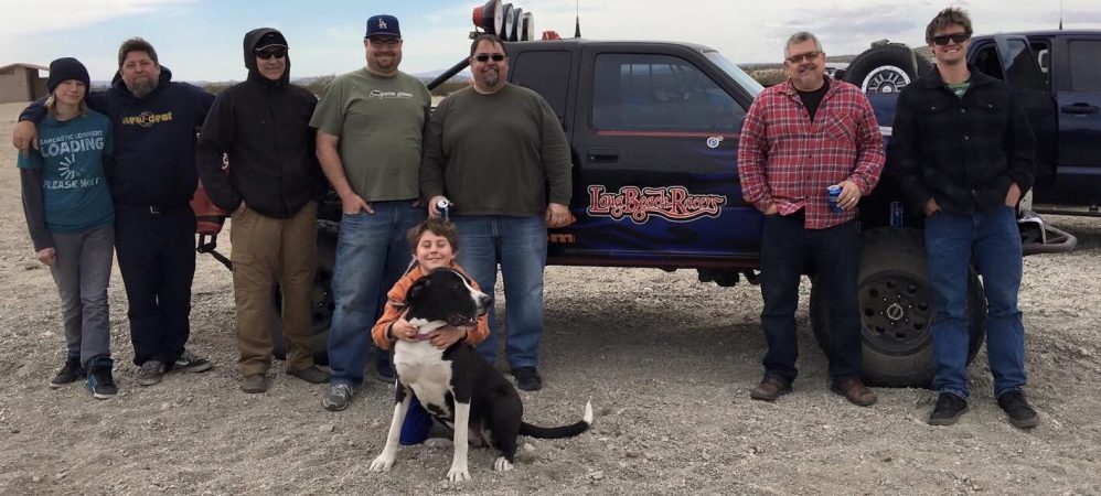 A group of seven adults and a child posing with a black dog in front of a Long Beach Racers off-road truck.