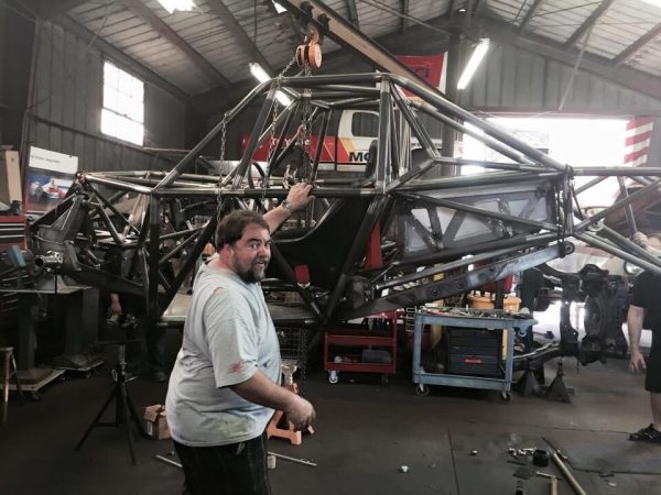 A man stands in a workshop next to a large, metal tube-frame chassis suspended by a crane.