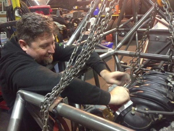 A man in a black sweatshirt works on the engine of a vehicle chassis suspended by heavy chains.